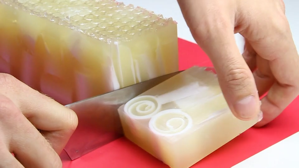 Hands slicing a translucent honey-toned melt-and-pour soap bar with a white spiral swirl in its center, on a red cutting board