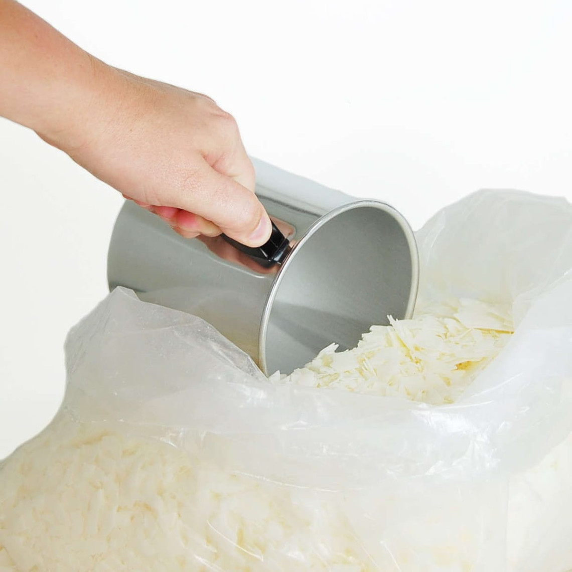 Hand scooping white soy wax flakes from a plastic bag with a silver pouring pitcher