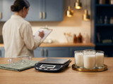 Cream candles, a kitchen scale, and safety glasses on a counter with a person checking a clipboard
