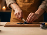 Maker in a mustard apron shaving a dark blue dye block with a knife on a cutting board, dye chips and candles nearby