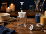 White wick-setter tool clamped over a clear glass jar centering a wick, beside a ceramic bowl, thermometer, wick spool, and blue, gold, and green candles