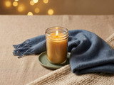 Lit amber candle in a glass jar on a sage dish atop a cream knit throw with a blue scarf and warm bokeh behind