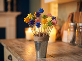 Colorful flower-shaped wax tarts on skewers in a rope-trimmed galvanized cup on a wooden counter by candlelight