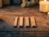 Four flat wooden wicks in metal tabs lined up on rustic wood beside lit jar candles