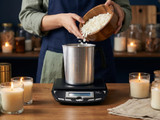 Hands pouring white soy wax flakes from a wooden bowl into a steel pitcher on a digital scale