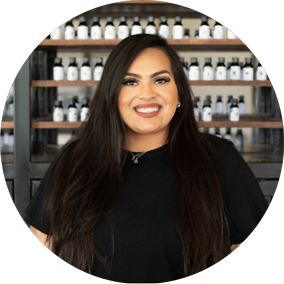 smiling woman with long dark hair in a black top before shelves of labeled black bottles
