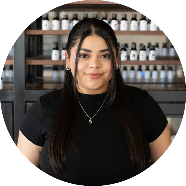 smiling woman with long straight dark hair and a pendant necklace before shelves of white bottles