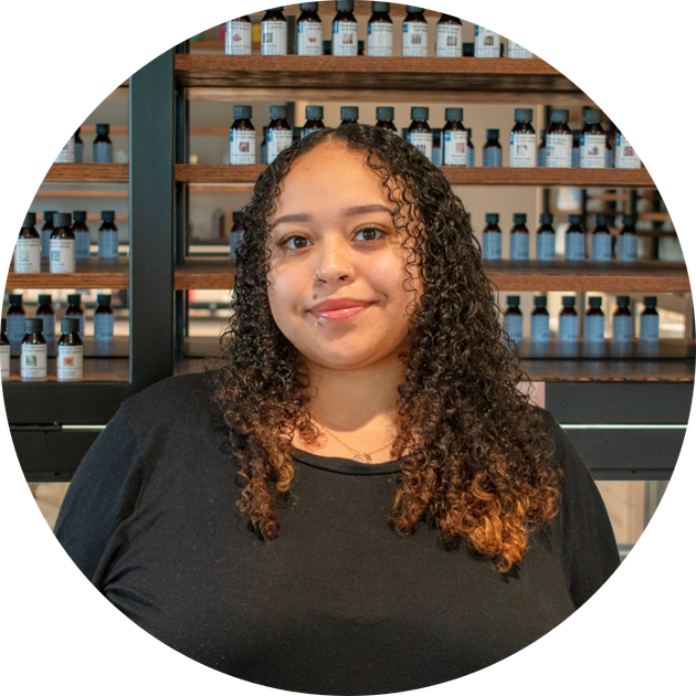 smiling woman with curly dark hair in a black top before wooden shelves of labeled bottles