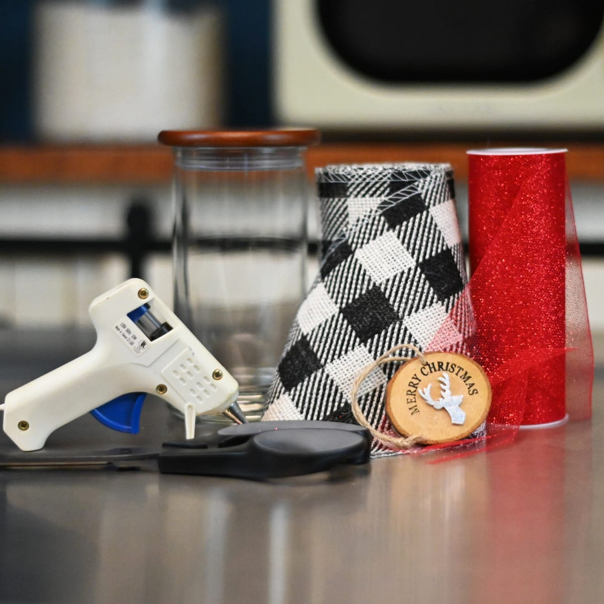 Glue gun, scissors, glass jar with wood lid, black-plaid and red sparkle ribbon rolls, and a Merry Christmas wood tag on a counter