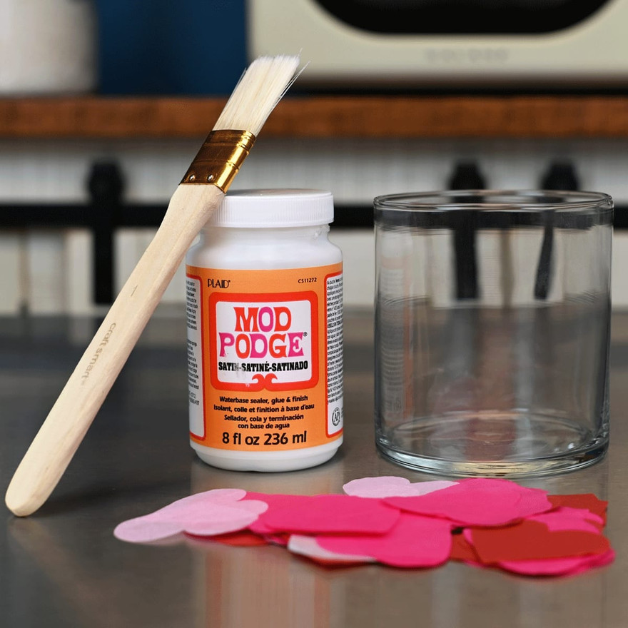 paintbrush resting on a bottle of Mod Podge next to a glass jar and a pile of pink and red tissue paper hearts