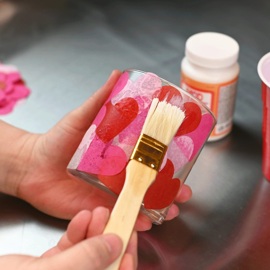 paintbrush applying a sealing coat over tissue paper hearts covering a glass jar