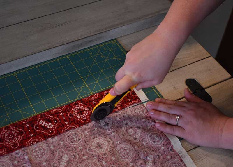 Hands cutting red paisley bandana fabric with a yellow rotary cutter on a green gridded cutting mat