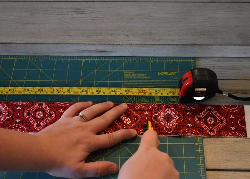 Hand holding red bandana strip flat as a rotary cutter trims it against a ruler on a green Olfa mat