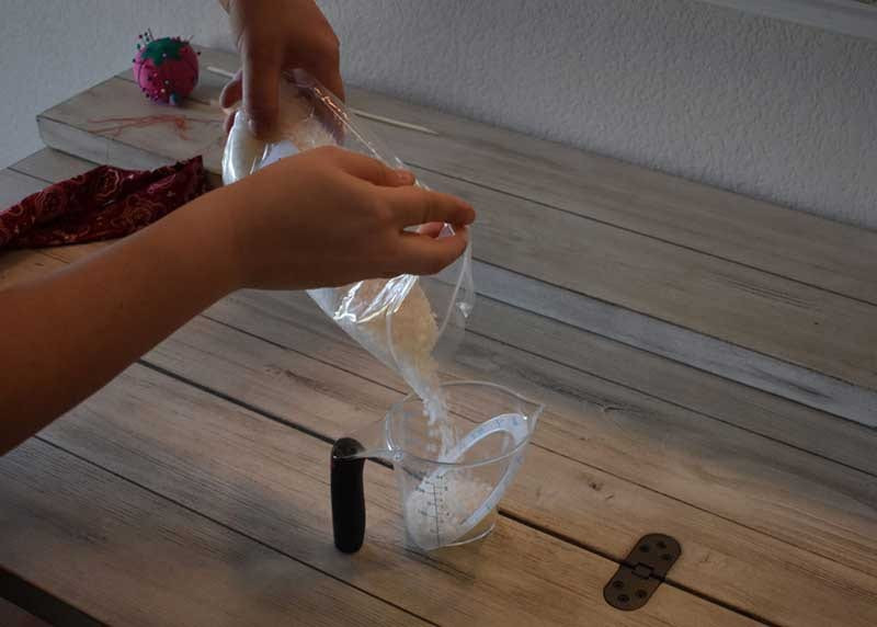 Hands pouring white water-absorbing crystals from a plastic bag into a clear measuring cup