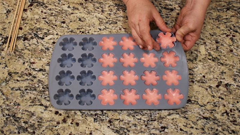 Hands easing a pink flower wax tart out of a gray silicone mold using a wooden skewer