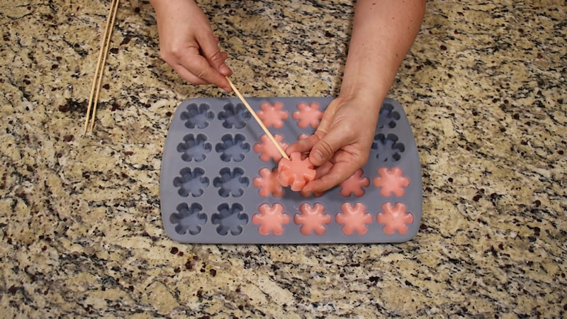 Hands pressing a wooden skewer into the back of a pink flower wax tart over a gray mold