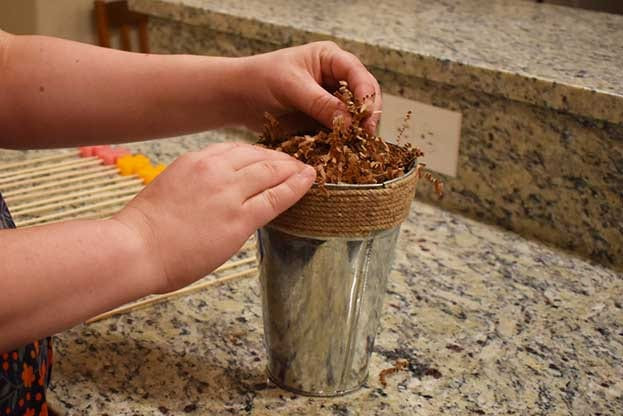 Hands tucking brown shredded paper into a rope-trimmed galvanized cup on a granite counter