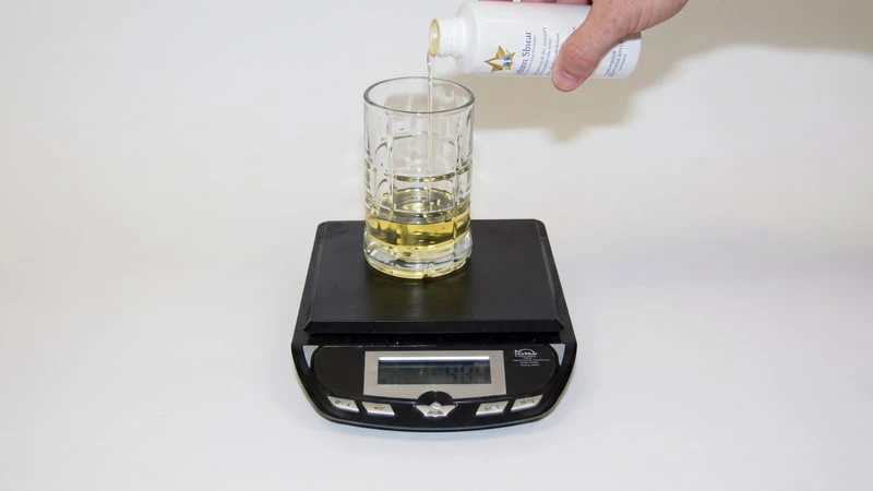 Hand pouring pale fragrance oil from a Lone Star Candle Supply bottle into a glass cup on a digital scale