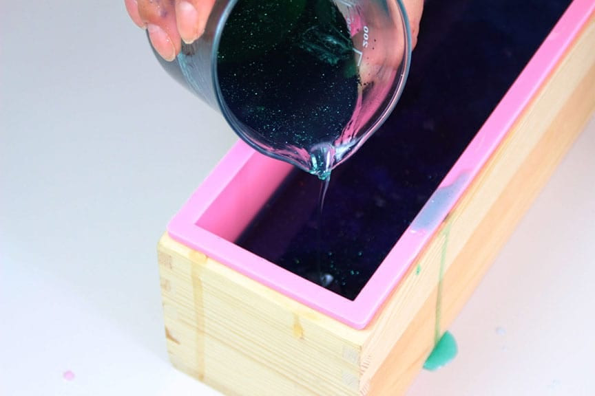 Dark teal glittery soap poured from a glass beaker into a pink silicone mold in a wooden frame