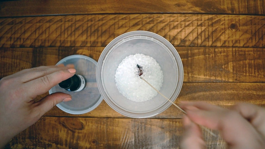 Wooden skewer stirring a drop of dark dye into white aroma beads in a clear tub, dye bottle on the lid