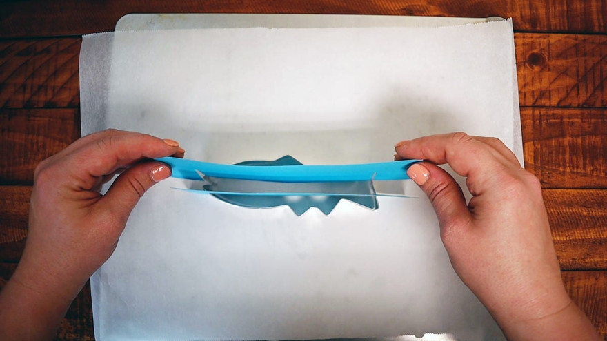Two hands bending a blue paper divider strip slotted lengthwise into a fish cookie cutter on parchment