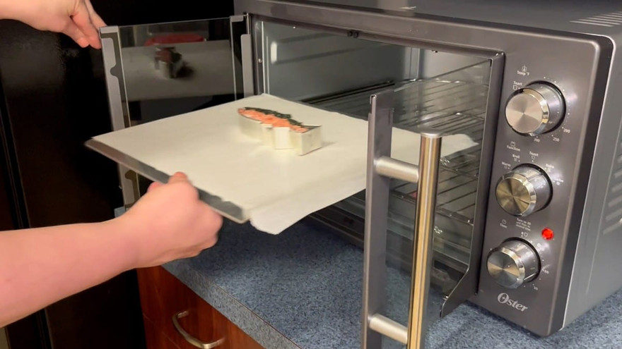 Hands sliding a parchment tray holding the filled fish cutter into a black Oster toaster oven