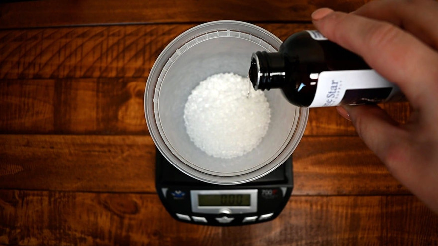 Hand pouring fragrance oil from a Lone Star amber bottle onto white aroma beads on a scale, overhead view