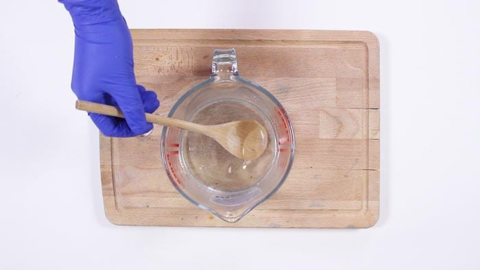 Blue-gloved hand stirring melted clear soap base with a wooden spoon in a glass jug