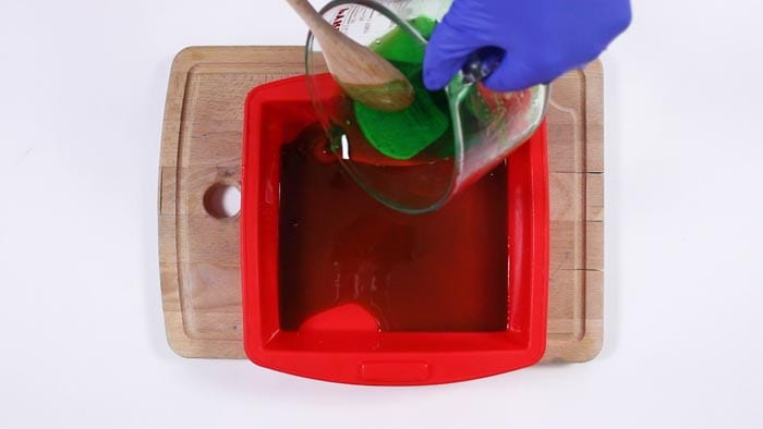 Blue-gloved hand pouring green melted soap from a jug into a red square silicone mold