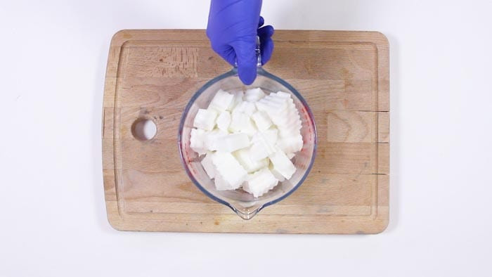 Blue-gloved hand steadying a glass jug of white soap base cubes on a wooden board
