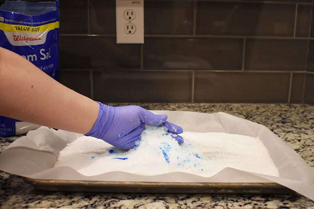 Purple-gloved hand mixing blue colorant into white Epsom salt on a parchment-lined sheet