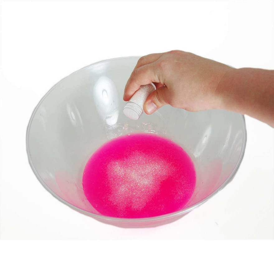 Hand shaking a vial of iridescent glitter over bright pink slime in a clear bowl