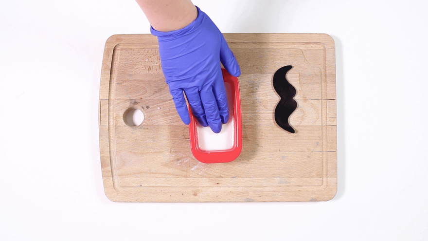 Blue-gloved hand pressing white soap into a small red mold, a black moustache shape on the board