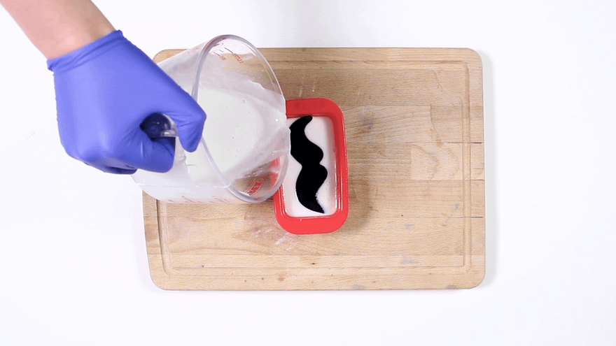Blue-gloved hand pouring white melted soap over a black moustache embed in a red rectangular mold