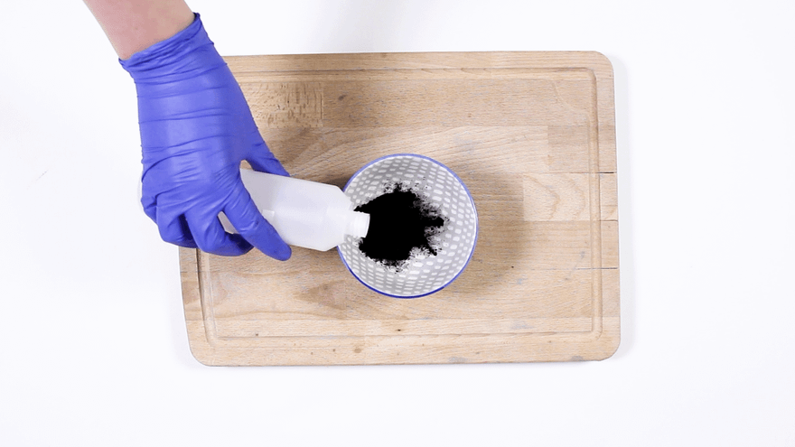 Blue-gloved hand pouring liquid from a white bottle onto black charcoal powder in a dotted bowl