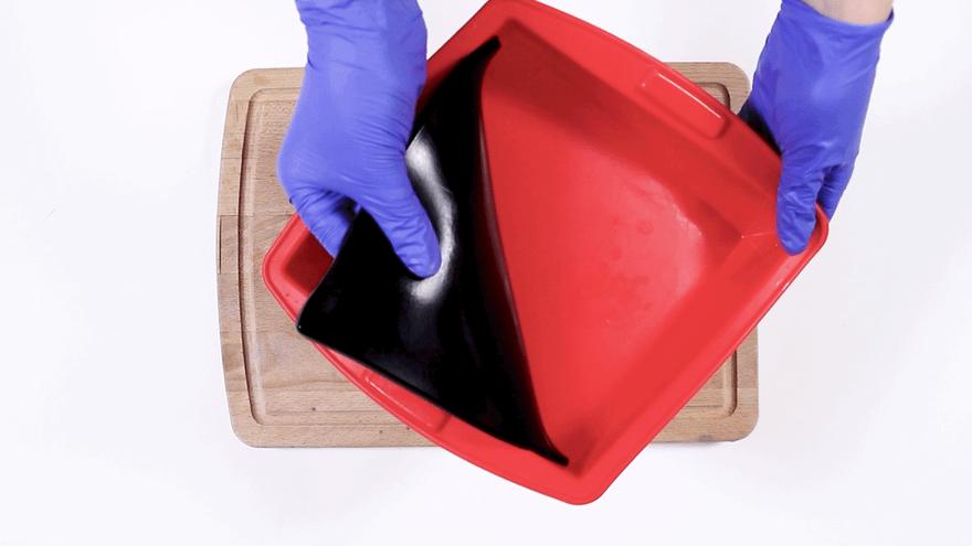 Blue-gloved hands peeling a set black soap slab out of a flexible red silicone mold