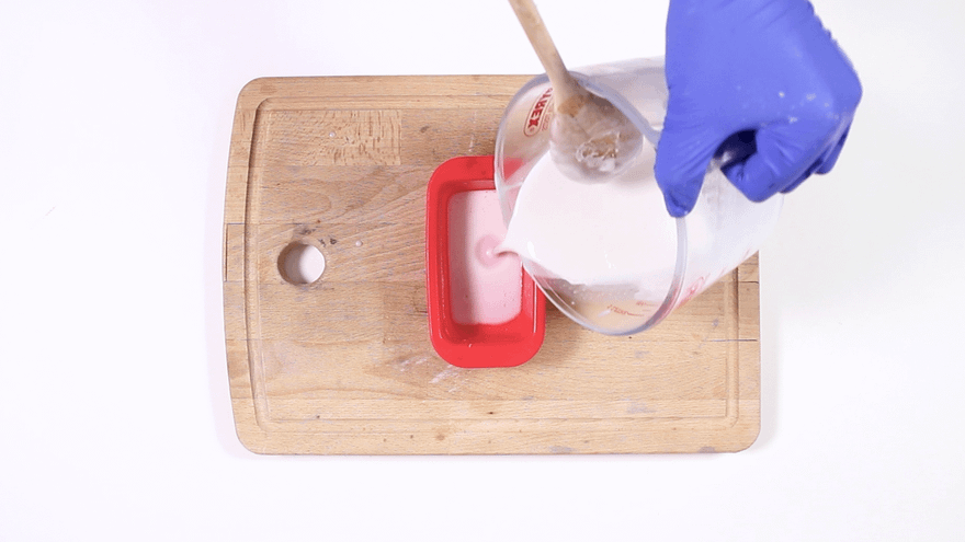 Blue-gloved hand pouring pink-tinted white melted soap into a small red rectangular silicone mold