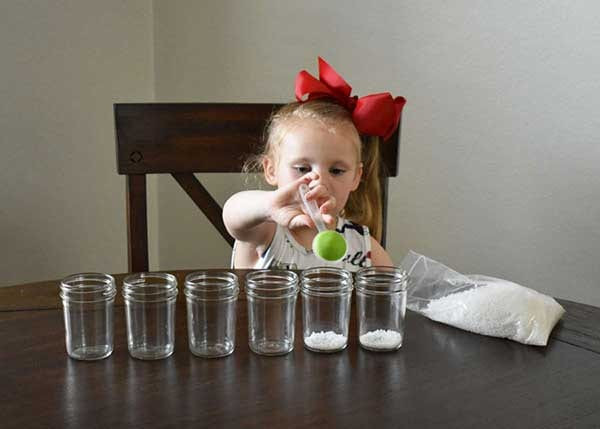 Young girl spooning white crystals into six glass jars beside an open bag of crystals
