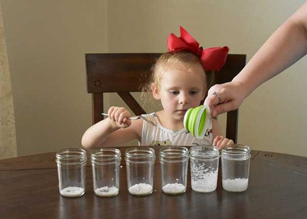 Adult tipping a green scoop of white crystals while a girl in a red bow spoons six mason jars