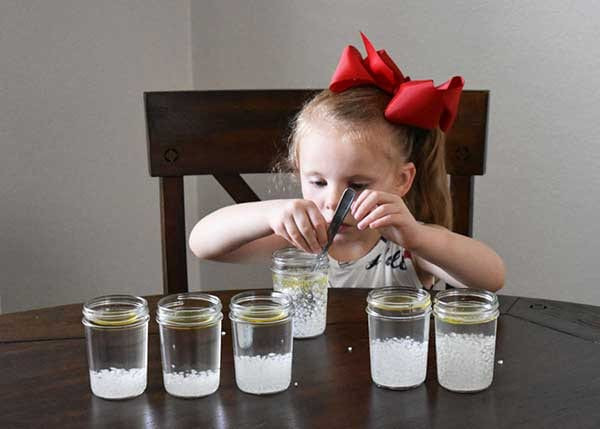 Girl in a red bow stirring water and white crystals in a mason jar, five jars lined up in front