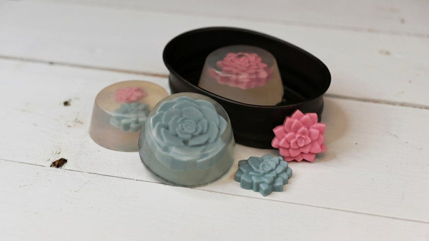 Teal and pink rose-shaped succulent soaps, some clear-cast, in and around a black oval tin