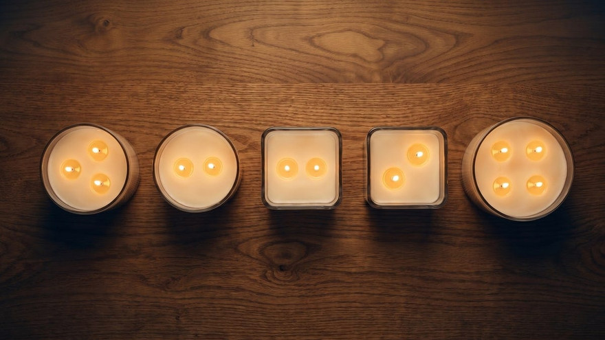 Overhead row of five lit multi-wick candles in round and square jars with two to four flames each