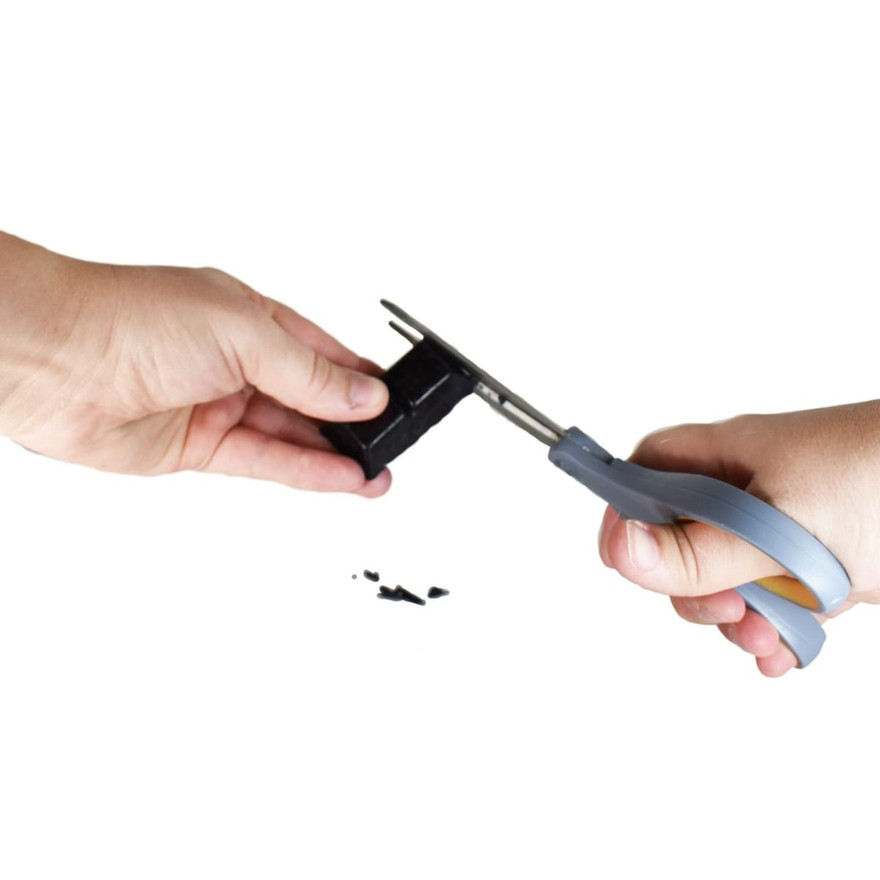 Two hands shaving a piece off a black dye block with gray scissors, small dye flakes below