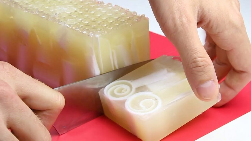 Hands slicing a bar from a translucent honey-toned melt-and-pour soap loaf on a red cutting board, the cut face showing two white spiral swirls and a bubble-textured top